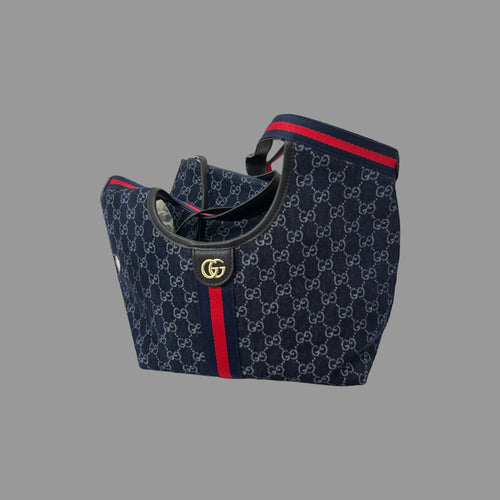 A navy blue Gucci tote bag featuring the brand's signature GG monogram pattern, red and navy web stripe, and a gold-tone interlocking GG logo on the front, set against a dark navy background.